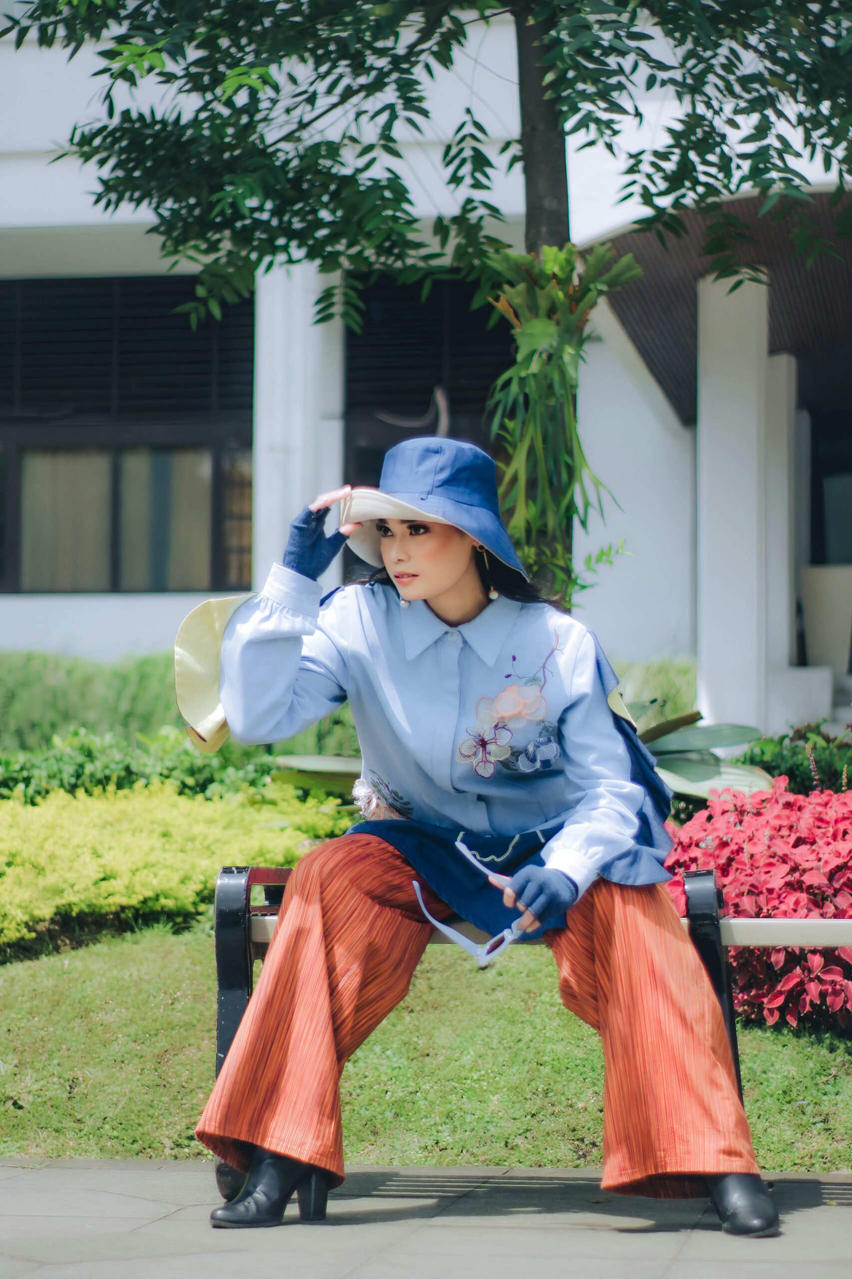 Stylish woman in vibrant outfit posing on a bench in a sunny Bandung park.
